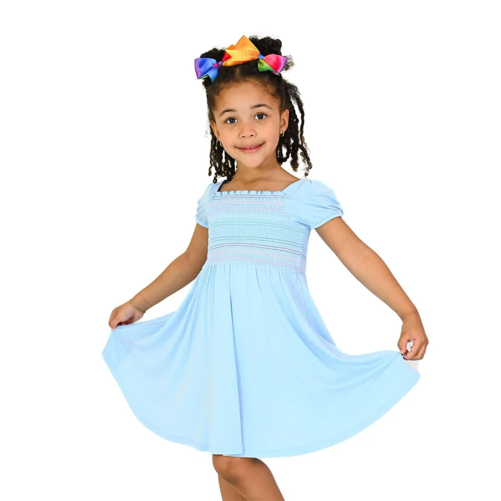 A young girl with braided hair wearing a light blue dress with smocked bodice details and a colorful bow in her hair, posing on a solid white background.