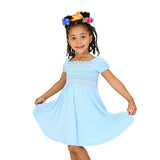 A young girl with braided hair wearing a light blue dress with smocked bodice details and a colorful bow in her hair, posing on a solid white background.