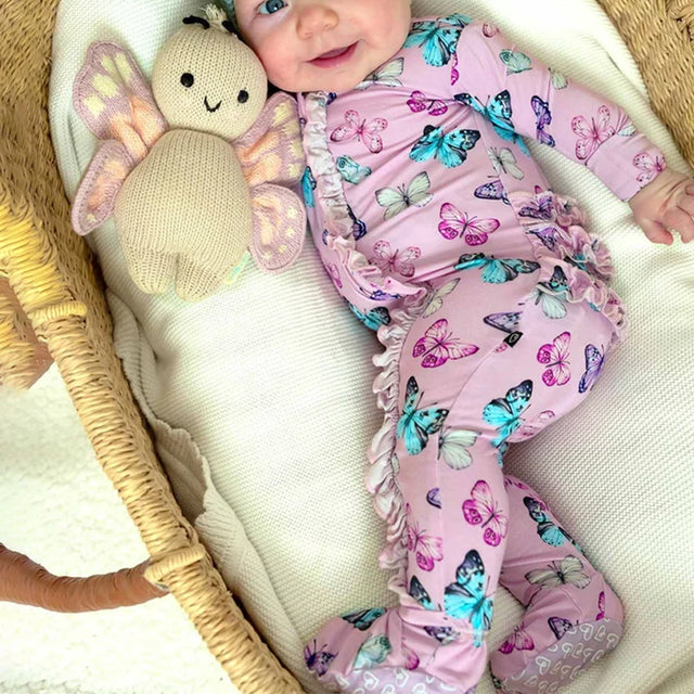 A baby wearing a pink footed sleeper with a colorful butterfly print and ruffled trim, lying in a woven basket bassinet next to a knitted butterfly toy.