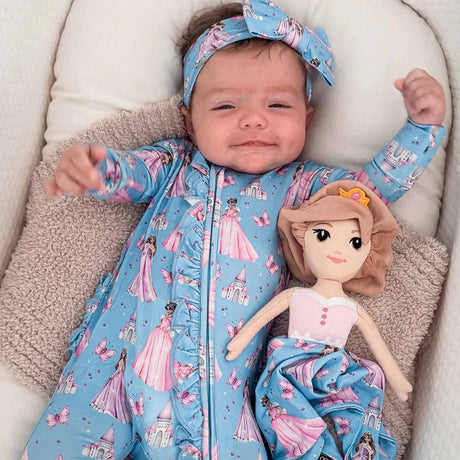 Baby in a blue outfit with princess designs holding a doll, lying on a beige cushion.