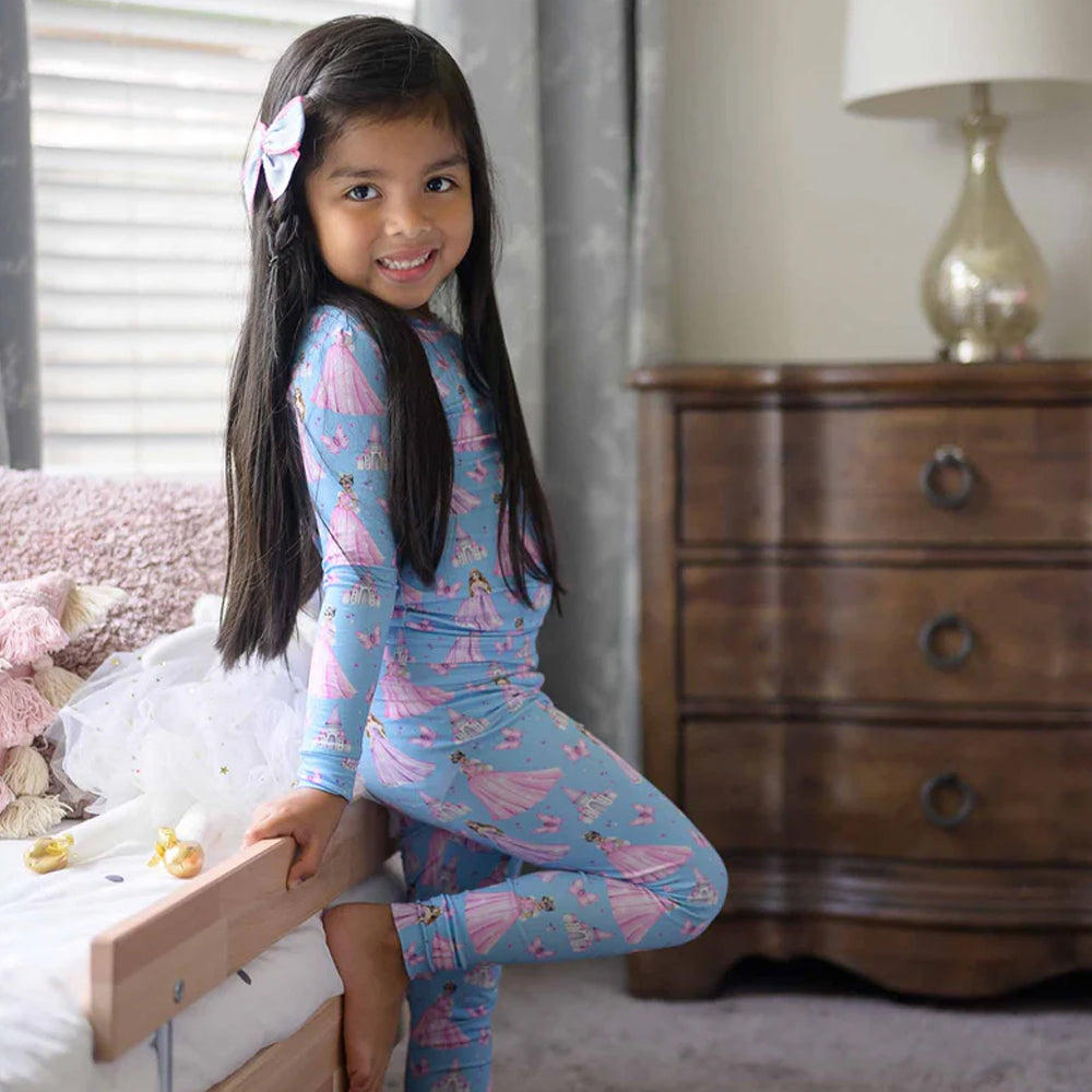 Young girl in blue pajamas with pink princess prints sitting on a bed in a bedroom.