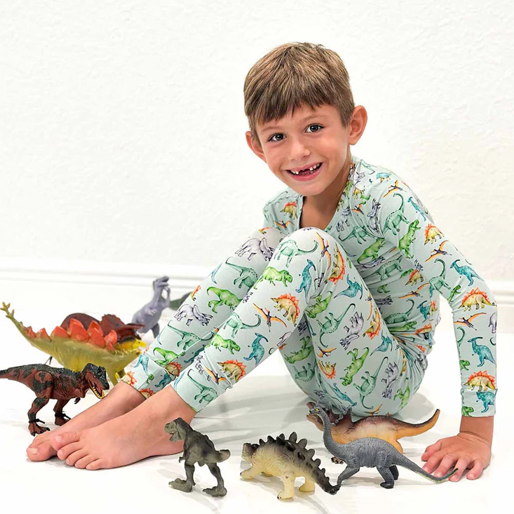 Child wearing pajamas with dinosaur pattern, sitting among toy dinosaurs on a white background