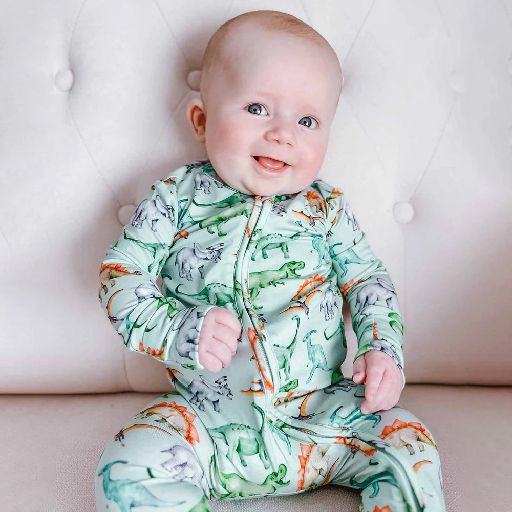 Baby wearing a dinosaur-patterned onesie sitting on a light-colored couch.