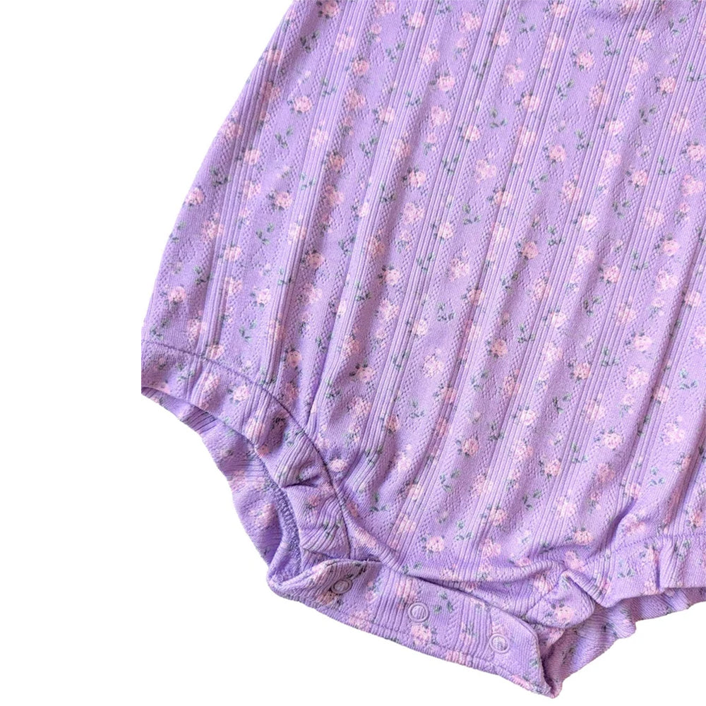 A close-up of a lavender ribbed fabric garment with a small pink and green floral print, showing the texture and snap-button closure at the bottom.