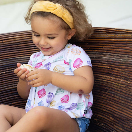 Child wearing a colorful macaron pattern outfit sitting on a wicker chair.