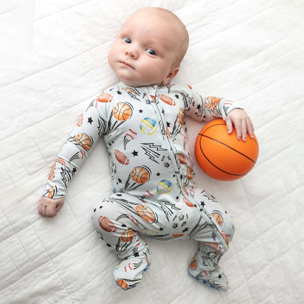 Baby wearing a onesie with sports-themed print, holding an orange ball on a white quilted surface.