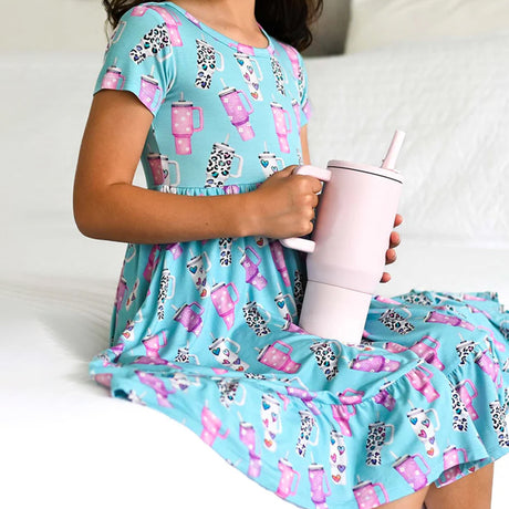 Child wearing a blue dress with pink and white cup print, holding a matching pink tumbler.