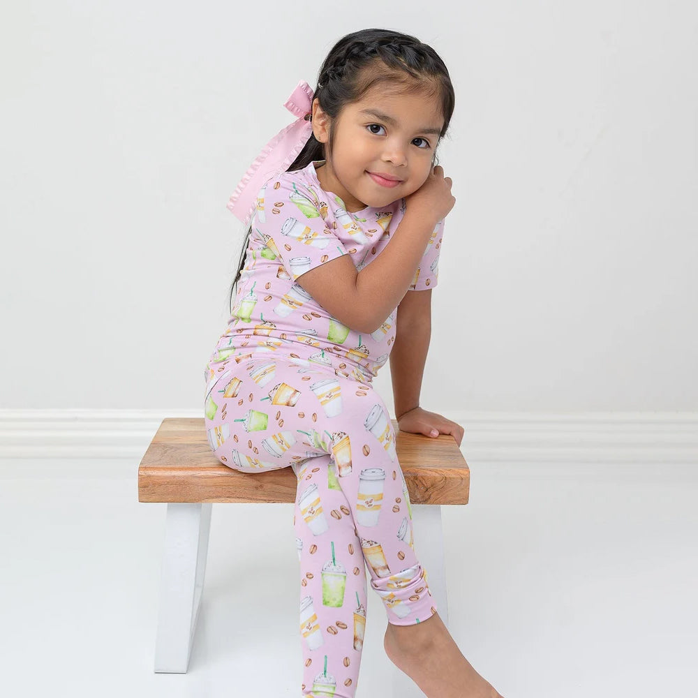 Young girl wearing a pink pajama set with cartoon designs, sitting on a wooden stool against a white background.