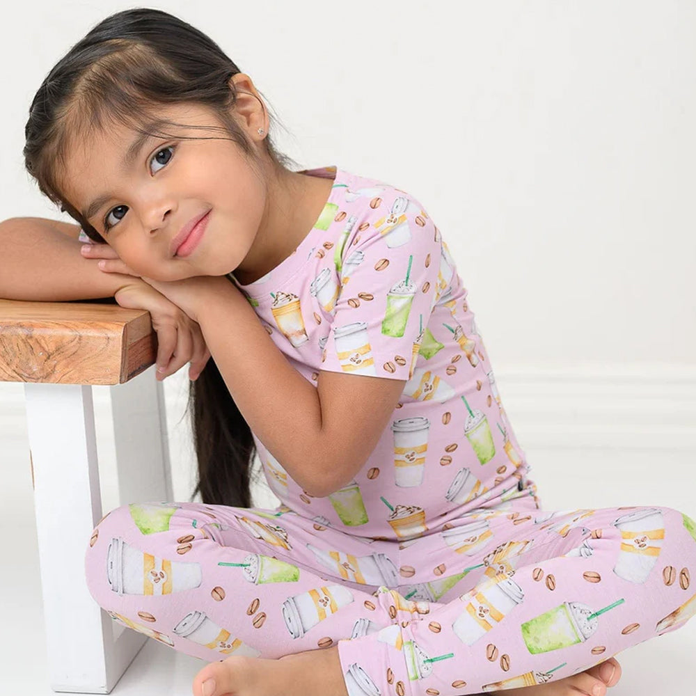 Young girl wearing pink pajamas with a drink pattern, sitting on a white surface.