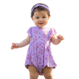 Baby in a purple floral romper standing outdoors with a tent in the background