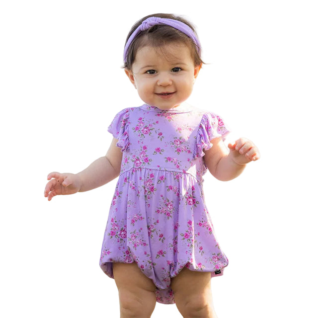 Baby in a purple floral romper standing outdoors with a tent in the background