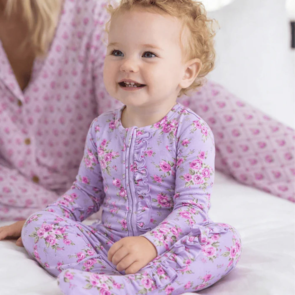 Child wearing a purple floral onesie sitting on a bed with a person in matching pajamas.