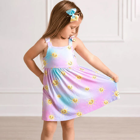 a girl wearing a dress with a pastel tie-dye gradient in pink, blue, and purple, featuring a yellow smiley sun print and a knotted shoulder strap, shown on a white background.