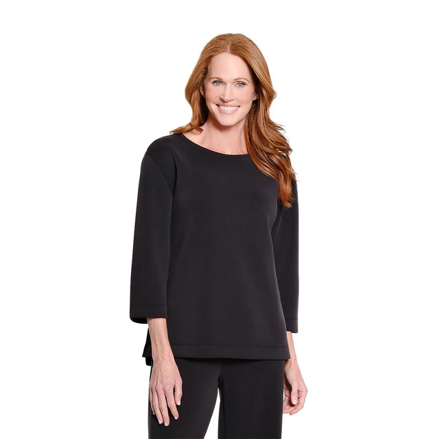 A smiling woman with long red hair wearing a black three-quarter sleeve top and black pants, standing against a plain white studio background.