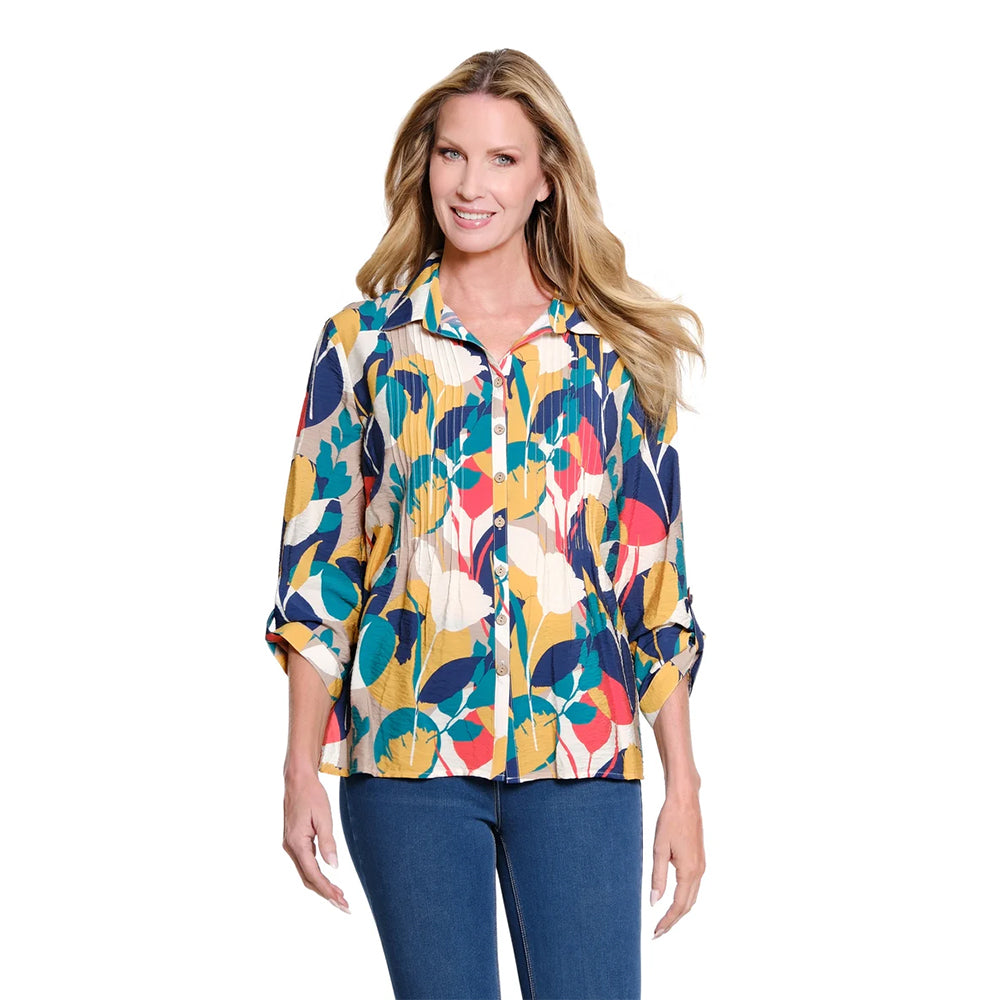 A woman modeling a colorful, long-sleeve pleated button-up blouse with a bold abstract floral pattern in yellow, teal, navy, red, and cream, paired with blue jeans against a solid white background.