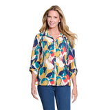 A woman modeling a colorful, long-sleeve pleated button-up blouse with a bold abstract floral pattern in yellow, teal, navy, red, and cream, paired with blue jeans against a solid white background.