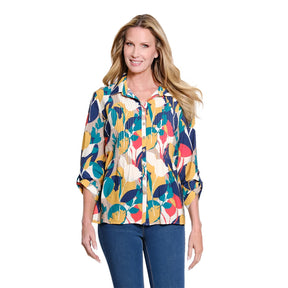 A woman modeling a colorful, long-sleeve pleated button-up blouse with a bold abstract floral pattern in yellow, teal, navy, red, and cream, paired with blue jeans against a solid white background.
