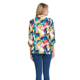 A woman modeling a colorful, long-sleeve pleated button-up blouse with a bold abstract floral pattern in yellow, teal, navy, red, and cream, paired with blue jeans against a solid white background.