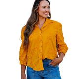 A woman wearing a mustard yellow lace button-up blouse and blue jeans, standing against a white wall.