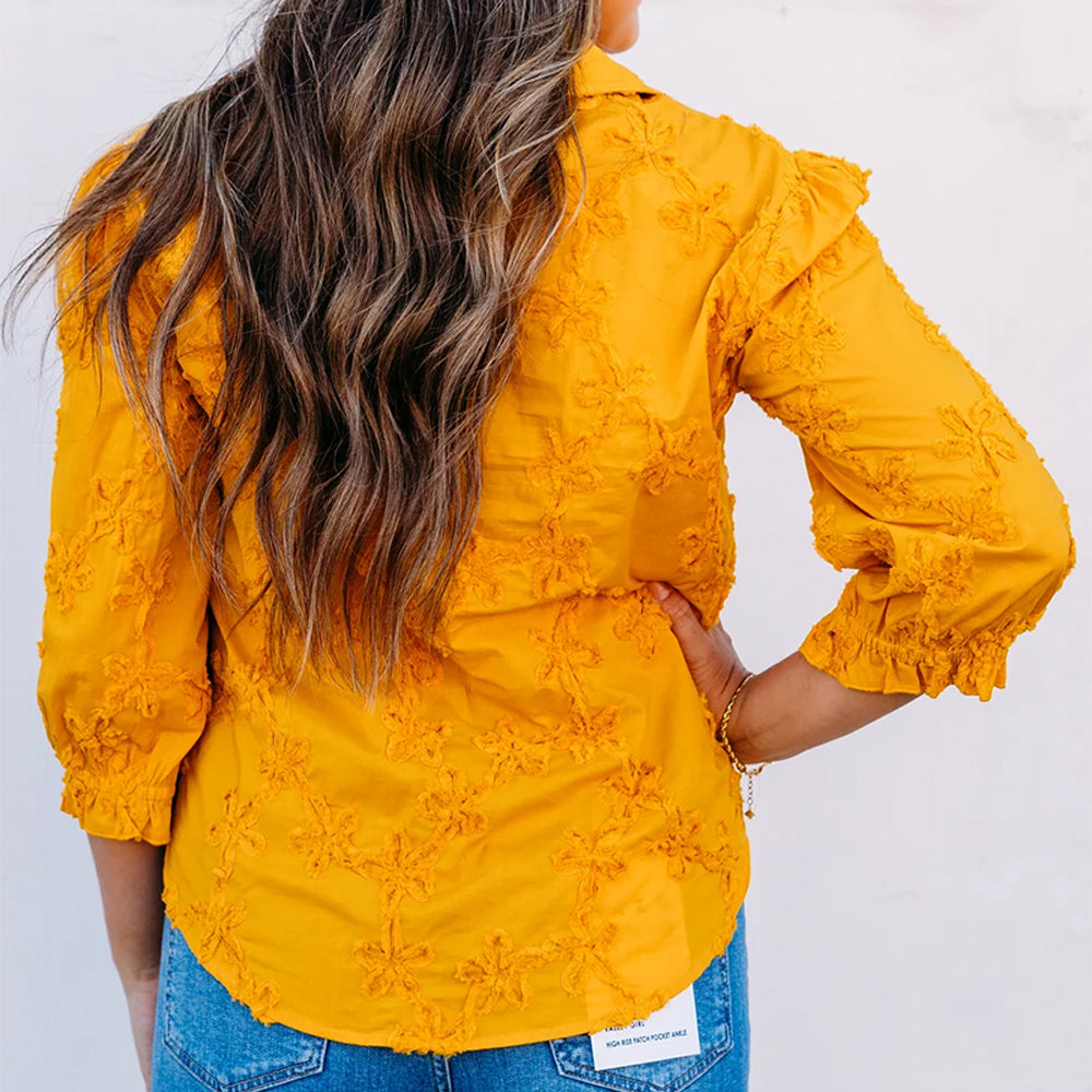 A woman viewed from the back wearing a mustard yellow long-sleeve blouse with floral embroidery and ruffled cuffs, paired with blue jeans against a plain white background.