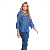 A woman modeling a blue denim button-up blouse with floral embroidery on the chest and three-quarter sleeves, paired with dark blue jeans against a plain white background.