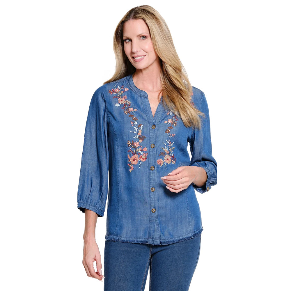 A woman modeling a blue denim button-up blouse with floral embroidery on the chest, paired with blue jeans, against a plain white background.