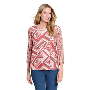 A woman modeling a three-quarter sleeve top with a geometric pink, red, and beige pattern, paired with blue jeans, against a plain white background.