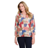 A woman modeling a long-sleeve collared blouse with a geometric patchwork pattern in beige, red, and blue, paired with dark navy pants, against a plain white background.