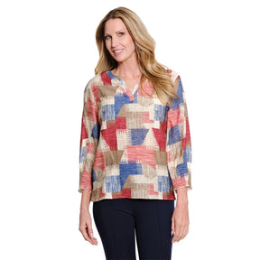 A woman modeling a long-sleeve collared blouse with a geometric patchwork pattern in beige, red, and blue, paired with dark navy pants, against a plain white background.