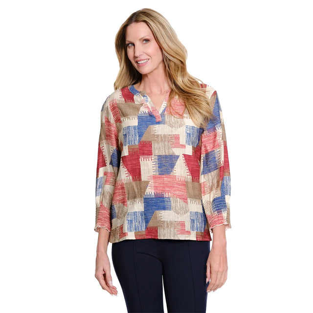 A woman modeling a long-sleeve collared blouse with a geometric patchwork pattern in beige, red, and blue, paired with dark navy pants, against a plain white background.