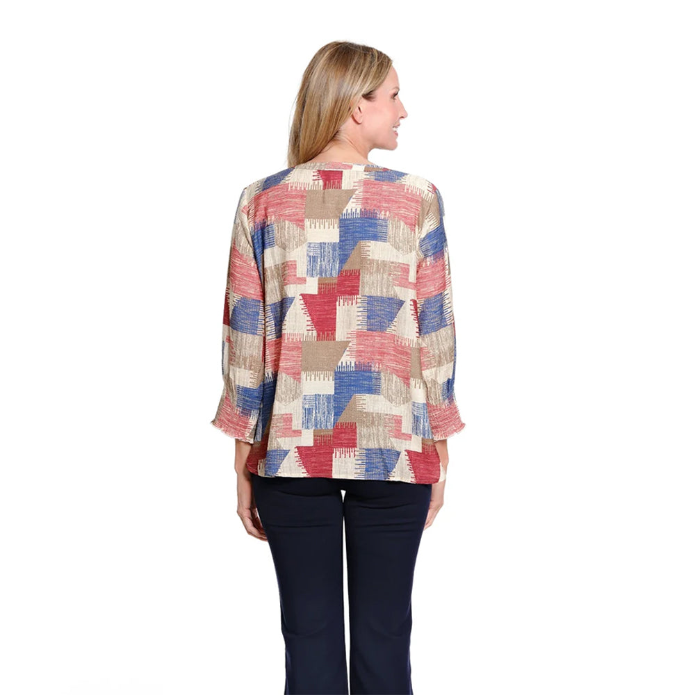 A woman viewed from the back wearing a long-sleeve blouse with a multicolor geometric patchwork pattern in red, blue, beige, and white, paired with dark navy pants, against a plain white background.