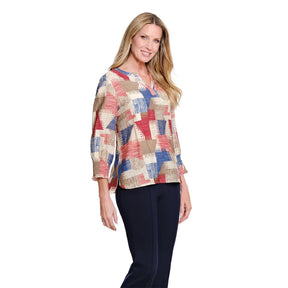 A woman modeling a three-quarter sleeve blouse with a multicolor geometric patchwork pattern in red, blue, beige, and brown, paired with dark navy pants, against a plain white background.