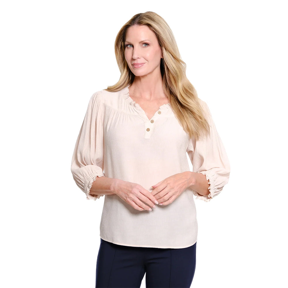 A woman modeling a light beige long-sleeve blouse with a ruffled V-neckline and three decorative buttons, paired with dark navy pants, against a solid white background.