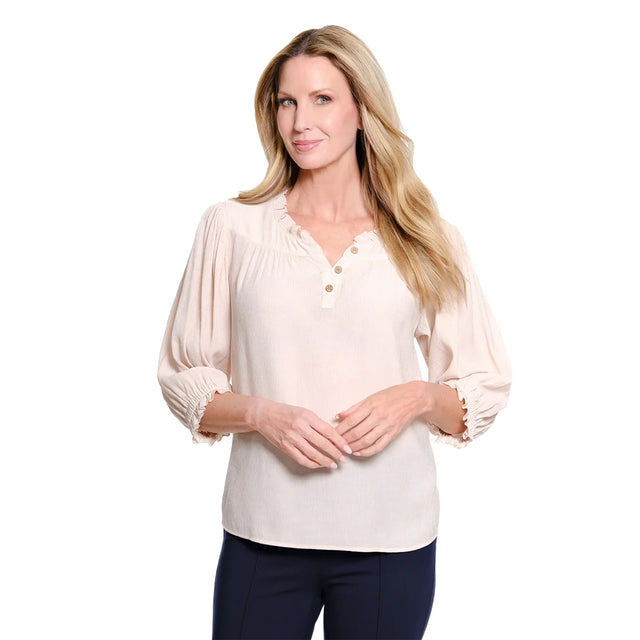 A woman modeling a light beige long-sleeve blouse with a ruffled V-neckline and three decorative buttons, paired with dark navy pants, against a solid white background.