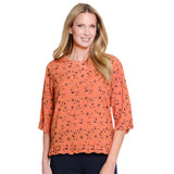 A woman modeling a short-sleeve orange top with a black abstract print, shown against a plain white background.