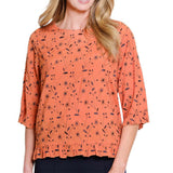 A woman wearing a peach-orange three-quarter sleeve blouse with a black floral and botanical print, featuring a ruffled hem and rolled cuffs, shown against a plain white background.
