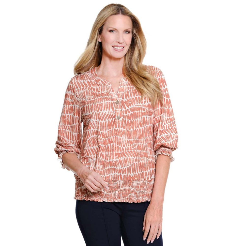 A woman modeling a long-sleeve blouse with a textured, abstract brushstroke pattern in terracotta orange and white, paired with dark pants, against a plain white background.