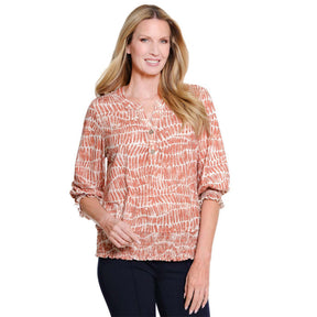A woman modeling a long-sleeve blouse with a textured, abstract brushstroke pattern in terracotta orange and white, paired with dark pants, against a plain white background.