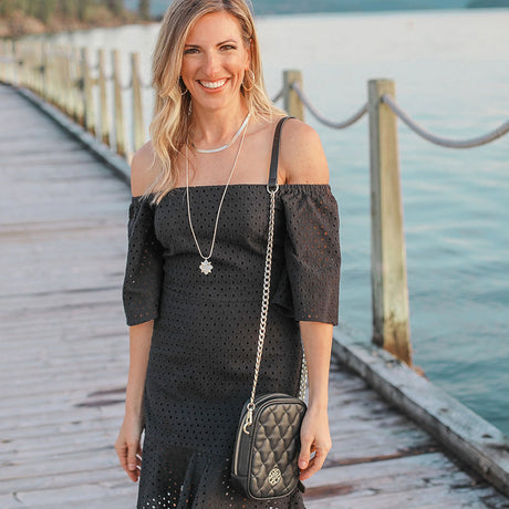 Woman in a black dress with a quilted handbag standing on a wooden dock by a body of water.