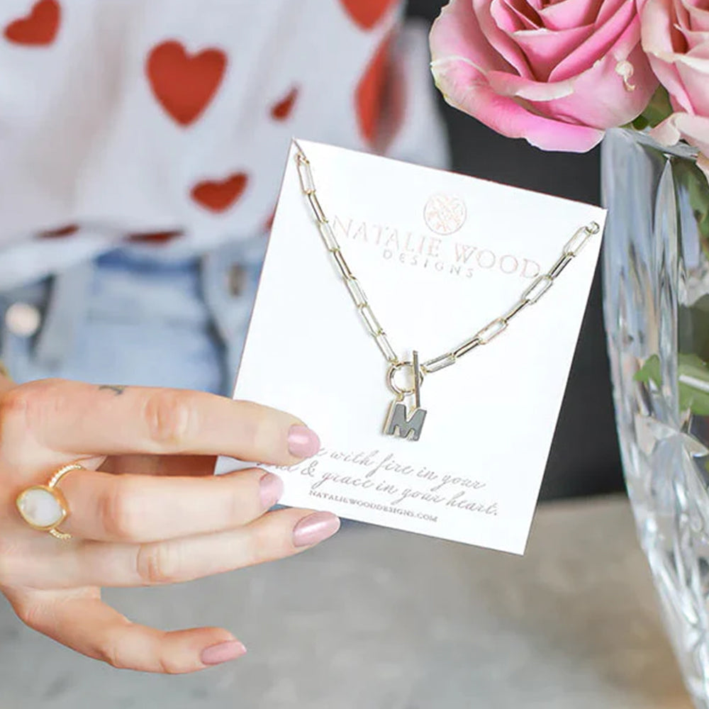 Gold necklace with 'IM' charm held by a hand on a card, with pink roses in the background.