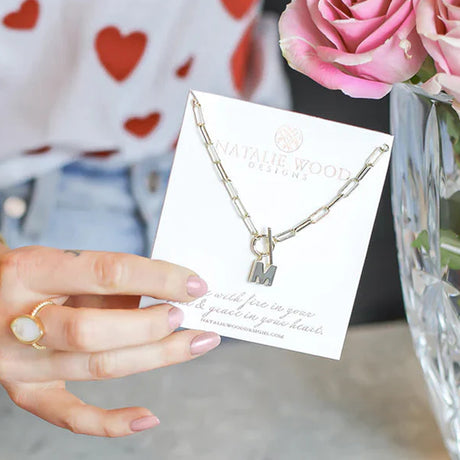 Gold necklace with 'IM' charm held by a hand on a card, with pink roses in the background.