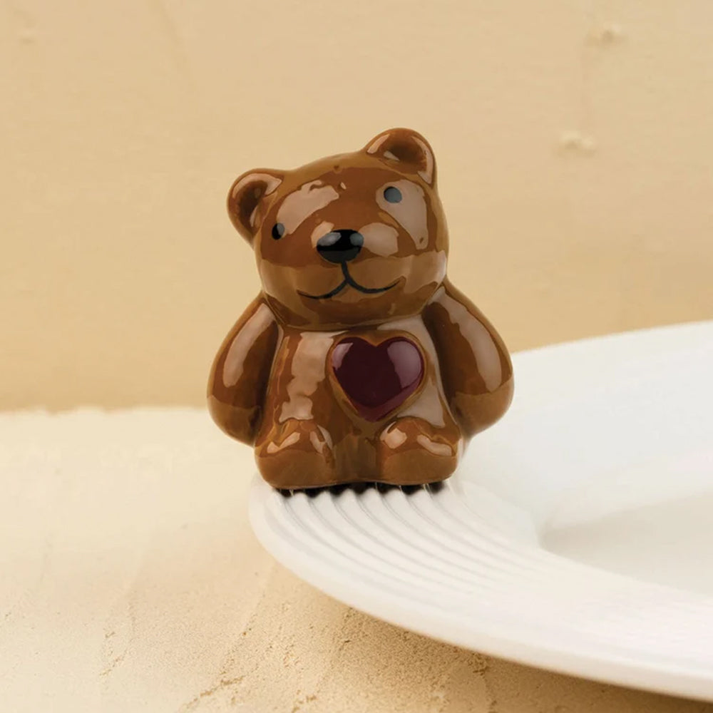 A glossy brown teddy bear-shaped object with a dark red heart on its chest, standing on a white plate against a warm beige background.