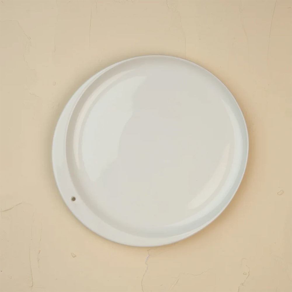 A plain white round wall-mounted plate or decorative disc with a small hole near the lower left edge, shown against a warm beige textured wall.