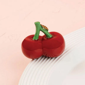 A glossy red cherry-shaped object with a green stem and small orange-and-black patterned leaf, photographed on a pale pink background with a white ribbed surface underneath.