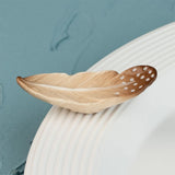 A decorative feather-shaped dish or spoon with a natural wood grain finish and white dotted details, resting on a white ribbed plate against a textured blue background.