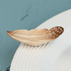 A decorative feather-shaped dish or spoon with a natural wood grain finish and white dotted details, resting on a white ribbed plate against a textured blue background.