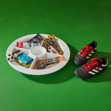 A white divided snack tray filled with assorted candy bars, a small soccer ball, and a cup, placed on a bright green background next to a pair of black soccer cleats with red laces and white stripes.