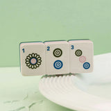Three white mahjong tiles with black, blue, and pink symbols resting on the edge of a white plate against a pale green background.