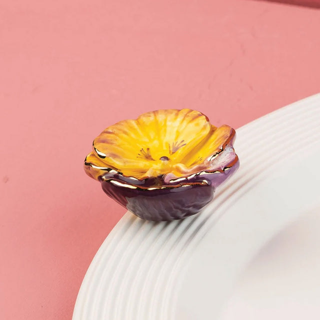 A small ceramic flower-shaped dish or candle holder with yellow and purple petals and gold trim, resting on a white ribbed plate against a pink background.