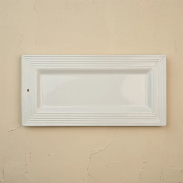 A white rectangular ceramic serving platter with a fluted border, shown against a textured beige wall.